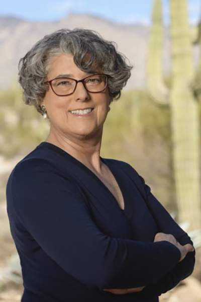 Caucasian woman wearing brown glasses and blue shirt with arms crossed in front of desert scene with saguaro cactus and mountains